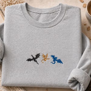 May include: A pale grey sweatshirt featuring three embroidered dragons in black, gold, and blue. The dragons are embroidered across the chest. The sweatshirt has a crew neck and long sleeves.