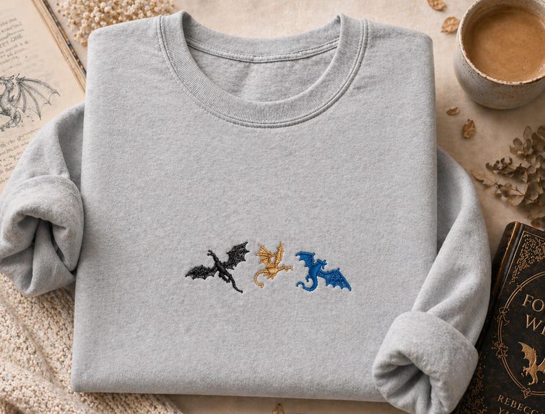 May include: A pale grey sweatshirt featuring three embroidered dragons in black, gold, and blue. The dragons are embroidered across the chest. The sweatshirt has a crew neck and long sleeves.