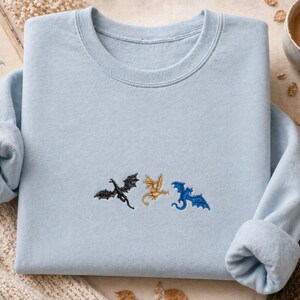 May include: A pale blue sweatshirt featuring three embroidered dragons in black, gold, and blue. The dragons are embroidered across the chest. The sweatshirt has a crew neck and long sleeves with rolled cuffs. A book and a cup of coffee are in the background.