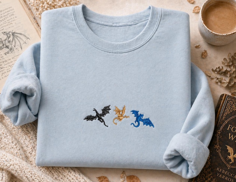 May include: A pale blue sweatshirt featuring three embroidered dragons in black, gold, and blue. The dragons are embroidered across the chest. The sweatshirt has a crew neck and long sleeves with rolled cuffs. A book and a cup of coffee are in the background.