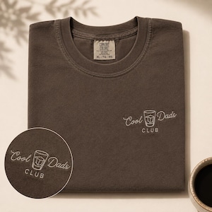 May include: A dark brown t-shirt with the embroidered text "Cool Dads Club" and a graphic of a cocktail glass. The shirt has a crew neck and a tag that reads "Comfort Colors XL-TG-EG".