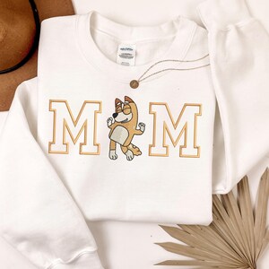 May include: A white long-sleeve crewneck sweatshirt featuring the embroidered text MOM in gold block letters, with a cartoon dog character replacing the letter O. The design is centered on the chest of the garment, which has a Gildan brand label.