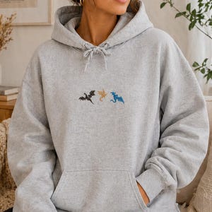 May include: A pale grey hooded sweatshirt with a drawstring and a front pocket. It has embroidered dragons in black, gold, and blue. The hoodie is made of a soft fabric.