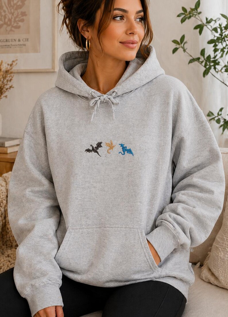 May include: A pale grey hooded sweatshirt with a drawstring and a front pocket. It has embroidered dragons in black, gold, and blue. The hoodie is made of a soft fabric.