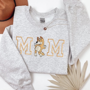 May include: A light gray crewneck sweatshirt featuring an embroidered design with the word MOM in gold block letters. The letter O is replaced by an embroidered illustration of a tan and white cartoon dog wearing sunglasses and dancing.