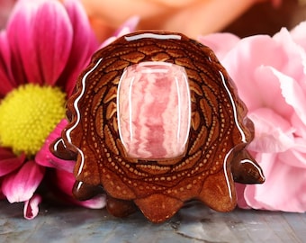 May include: A handcrafted wooden pendant with a central, rectangular pink gemstone. The pendant is surrounded by intricate wood grain patterns and rests on a surface with a floral arrangement of pink and peach flowers.