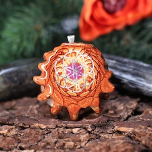 Puede incluir: Un colgante de madera con un diseño geométrico plateado y rosa. El colgante tiene forma de flor y tiene una anilla plateada.