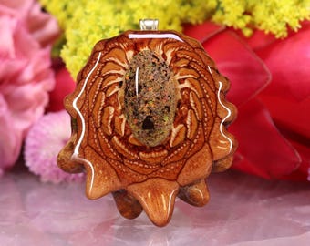 May include: A handcrafted pendant with a central textured stone set in a detailed, organic-shaped wooden frame. The pendant has warm brown tones with intricate patterns, and a silver bail for a necklace. The background includes colorful flowers.