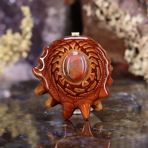 Puede incluir: Colgante artesanal con una piedra preciosa central ovalada, de color marrón rojizo, engastada en una base de madera tallada detallada. La base de madera es de color marrón cálido con patrones intrincados y una argolla de metal.