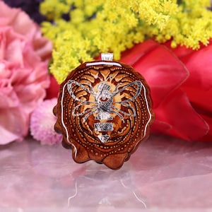 Puede incluir: Un colgante marrón con un diseño de abeja plateada. El colgante es redondo y tiene una superficie texturizada. El diseño de la abeja está detallado con líneas intrincadas. El colgante se muestra sobre un fondo de flores coloridas.