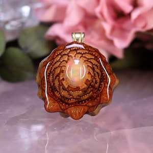 Puede incluir: Un colgante artesanal con un diseño natural de piña de color marrón claro y una fianza dorada. El colgante tiene una piedra preciosa de ópalo ovalada central. El fondo incluye flores rosas y hojas verdes.