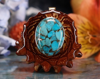 May include: A handcrafted pendant featuring a vibrant turquoise stone with gold flecks, set in a detailed, dark brown wooden frame. The pendant has a silver bail and is designed to be worn as a necklace. The wooden frame has a unique, textured design.
