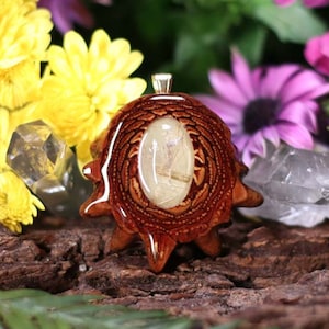 Puede incluir: Un colgante de resina marrón y puntiagudo con un cristal de cuarzo transparente en su interior. El colgante está rodeado de flores.