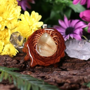 Puede incluir: Un colgante de madera marrón con un cristal de cuarzo transparente encapsulado en resina. El colgante tiene un diseño detallado e intrincado y está rodeado de flores.