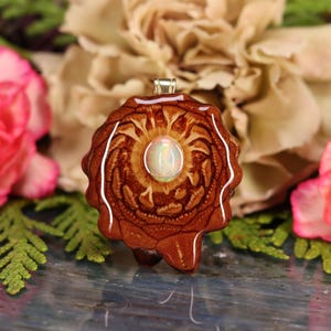 Puede incluir: Un colgante marrón y dorado con un diseño circular y una piedra de ópalo blanca en el centro.