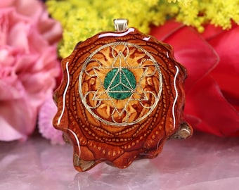May include: A handcrafted wooden pendant with a detailed, intricate design. The pendant features a central green stone, surrounded by a gold-colored wire design and set within a brown wooden base. The pendant is a unique piece of jewelry.