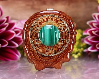 Può includere: Ciondolo artigianale con una pietra di malachite verde lucida incastonata in un dettagliato design a filigrana dorata. Il ciondolo è racchiuso in una base di legno marrone intagliato, che mostra motivi naturali delle venature del legno. Design elegante.