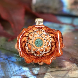 Puede incluir: Un colgante de madera con un diseño de metal dorado y una piedra turquesa en el centro. El colgante tiene forma de flor y presenta una gama de colores marrón y naranja.