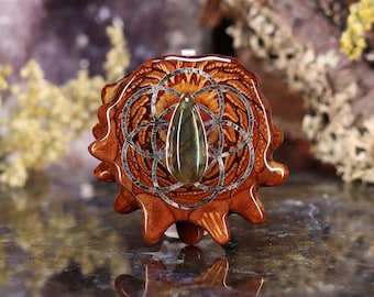 May include: A handcrafted pendant featuring a central, teardrop-shaped gemstone set within a silver-toned, geometric design. The pendant is encased in a polished, reddish-brown wooden base with a unique, textured pattern. The overall design is intricate and artistic.