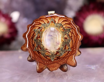 May include: A handcrafted pendant featuring a central oval-shaped, iridescent gemstone. The stone is set within a gold-colored, star-shaped frame, surrounded by a detailed, brown, wooden base. The pendant has a small, gold-colored bail for a chain.