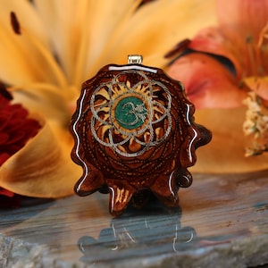 Puede incluir: Un colgante de madera marrón con un diseño de mandala plateado y dorado y una piedra preciosa verde en el centro. El colgante presenta el símbolo Om en el centro de la piedra preciosa.
