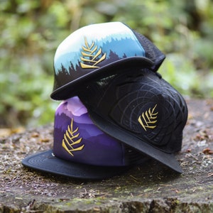Pode incluir: Dois bonés de caminhoneiro empilhados um em cima do outro. O boné de cima é azul com uma cena de montanha branca e verde e um design de samambaia dourado. O boné de baixo é roxo com uma cena de montanha branca e rosa e um design de samambaia dourado. Ambos os bonés têm costas de malha preta.