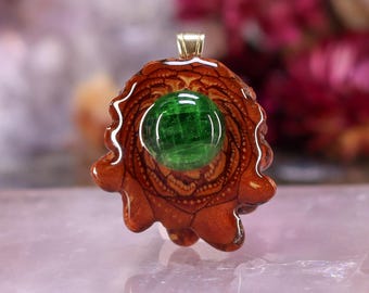 May include: A handcrafted pendant featuring a vibrant green gemstone set in a detailed, reddish-brown wooden base. The pendant has a gold-colored bail for a necklace chain. The wood has a unique, organic shape.