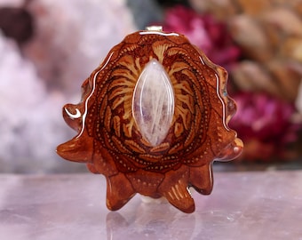 May include: A handcrafted pendant featuring a central, oval-shaped, translucent stone set within a detailed, brown, wooden base. The wood has intricate patterns and a natural, organic shape. The pendant has a small, silver-colored bail.