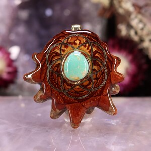 Puede incluir: Colgante artesanal con una piedra turquesa engastada en un marco de metal dorado. El colgante tiene una forma única con un diseño detallado en tonos marrón y beige. El colgante mide aproximadamente 5 cm de diámetro.