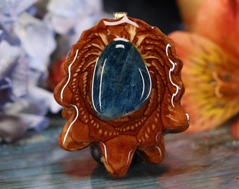Op de afbeelding: Handgemaakte hanger met een gepolijste, ovaalvormige blauwe edelsteen gezet in een gedetailleerde, bruine houten lijst. De hanger heeft een goudkleurige balein voor een ketting. Het hout heeft een natuurlijk, organisch ontwerp.