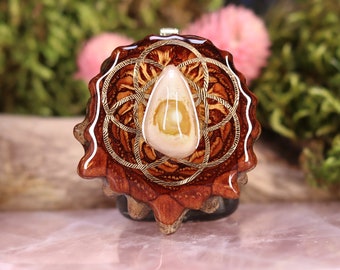 May include: A brown wooden pendant with a gold-colored geometric design and a white teardrop-shaped stone in the center.