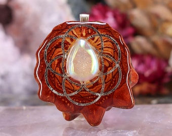 May include: A handcrafted pendant featuring a teardrop-shaped, iridescent crystal at the center. The crystal is set within a detailed, silver-colored geometric design, surrounded by a reddish-brown, organic-shaped wooden base. The pendant has a silver bail.