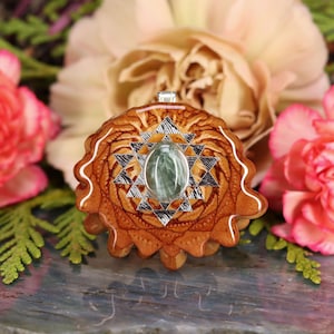 Puede incluir: Un colgante de madera con un diseño geométrico plateado y una piedra preciosa verde en el centro.