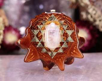 May include: A handcrafted pendant featuring a central oval-shaped, translucent stone. The pendant is set within a geometric design of gold and brown tones, with a natural wood-like texture. The overall shape is organic and unique.