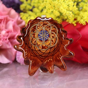 Puede incluir: Colgante marrón con un diseño intrincado. Presenta un patrón geométrico dorado sobre un fondo azul y beige. El colgante es de madera y tiene una anilla plateada. El diseño del colgante es detallado.