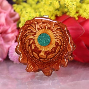 Puede incluir: Colgante artesanal con un diseño único. Presenta una piedra central de color turquesa rodeada de un patrón de madera tallada detallado en tonos marrones y beige. El colgante tiene una argolla plateada para colgar.