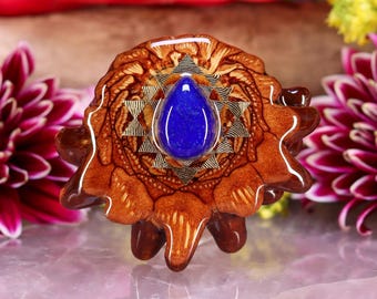 May include: A handcrafted pendant featuring a teardrop-shaped, deep blue gemstone set in a starburst design. The gemstone is surrounded by a detailed, brown, wooden base with a unique, organic shape. The pendant is displayed on a reflective surface with floral accents.