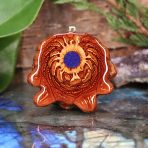 Puede incluir: Un colgante de madera con una piedra de lapislázuli azul en el centro. La madera es de color marrón rojizo y tiene un patrón de espiral. El colgante tiene forma de flor.