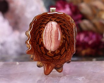 May include: A handcrafted pendant featuring a central oval-shaped pink gemstone with white stripes, set within a detailed, brown, wood-like resin. The pendant has a gold-colored bail for a necklace.