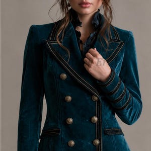 Puede incluir: Un blazer cruzado de terciopelo color verde azulado con ribetes de terciopelo negro en las solapas, pu&ntilde;os y bolsillos. La chaqueta est&aacute; adornada con botones dorados texturizados dispuestos en dos filas verticales y un cuello con volantes.