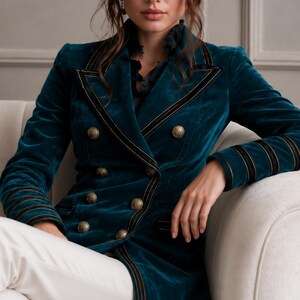 Puede incluir: Un blazer cruzado de terciopelo color verde azulado con ribetes negros y dorados en las solapas y pu&ntilde;os, junto con seis botones met&aacute;licos redondos. La chaqueta tiene un cuello estructurado y mangas largas, sobre una camisa oscura con volantes.