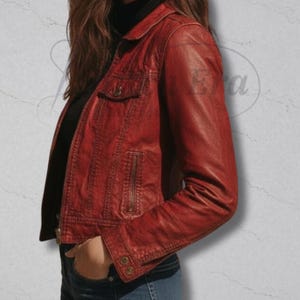 May include: A red leather jacket with a collared neckline, a small chest pocket, and a zippered front. The jacket has buttoned cuffs and a tailored fit. The model is wearing a black turtleneck underneath. The jacket is a deep shade of red.