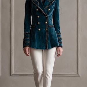 Puede incluir: Un blazer cruzado de terciopelo color verde azulado con botones dorados, ribetes negros en las solapas y pu&ntilde;os, y un corte estructurado, combinado con pantalones ajustados de color blanco roto y zapatos de tac&oacute;n neutros.