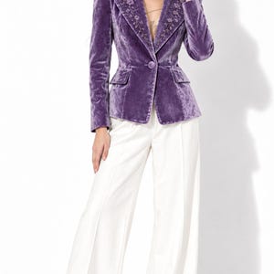 Puede incluir: Una americana de terciopelo morado con cierre de un solo botón, hombros estructurados y delicados bordados florales en las solapas, combinada con pantalones blancos de talle alto y pernera ancha con una raya central marcada.