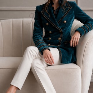 Puede incluir: Un blazer cruzado de terciopelo color verde azulado con botones dorados, ribetes negros en las solapas y pu&ntilde;os, combinado con pantalones blancos ajustados y zapatos de tac&oacute;n alto en color dorado metalizado. El blazer tiene un corte estructurado.