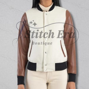 Puede incluir: Chaqueta universitaria color crema con mangas de cuero marrón. La chaqueta tiene cuello, puños y dobladillo de canalé negro. Cuenta con cierre de botones y dos bolsillos delanteros. El texto "Stitch Era Boutique" está impreso en la chaqueta.