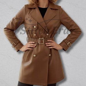 May include: A brown trench coat with a double-breasted design, featuring gold-coloured buttons and a matching belt with a square buckle. The coat has a classic collar and long sleeves with buckle details at the cuffs. The text "Stitch Era Boutique" is visible.