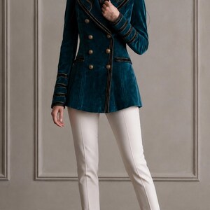 Puede incluir: Un blazer cruzado de terciopelo color verde azulado con ribetes negros, botones dorados y hombros estructurados, combinado con pantalones color crema de corte ajustado y tacones de punta. El blazer tiene mangas largas con bandas negras.
