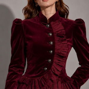 May include: A deep burgundy velvet jacket featuring a high collar, long sleeves with ruffled cuffs, and a row of metallic buttons. The front is accented with a vertical pleated ruffle detail that extends into a layered peplum hem at the waist.