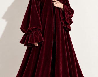 May include: A floor-length, deep burgundy velvet cloak featuring a hood with a ruffled edge, long bell sleeves with ruffled cuffs, and a ruffled hemline. The garment includes a tie closure at the neckline and a full, flowing silhouette.